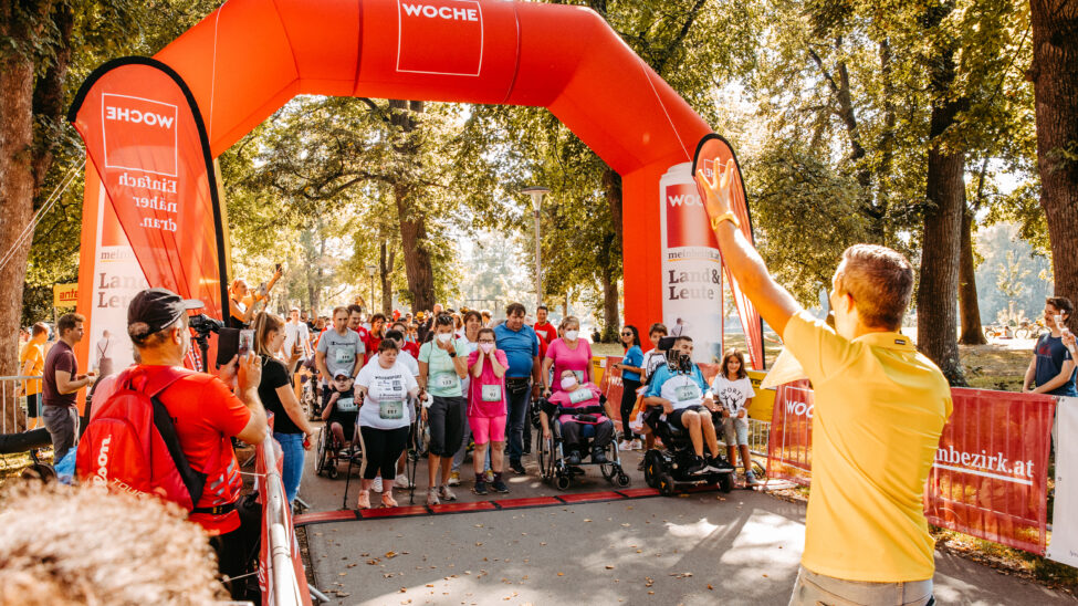 „Startbereich eines inklusiven Laufs im Park: Menschen mit und ohne Behinderung stehen unter einem großen roten Startbogen. Einige sitzen im Rollstuhl, andere stehen daneben. Ein Mann im gelben Shirt gibt ein Startsignal, während Zuschauende am Rand applaudieren und fotografieren.“