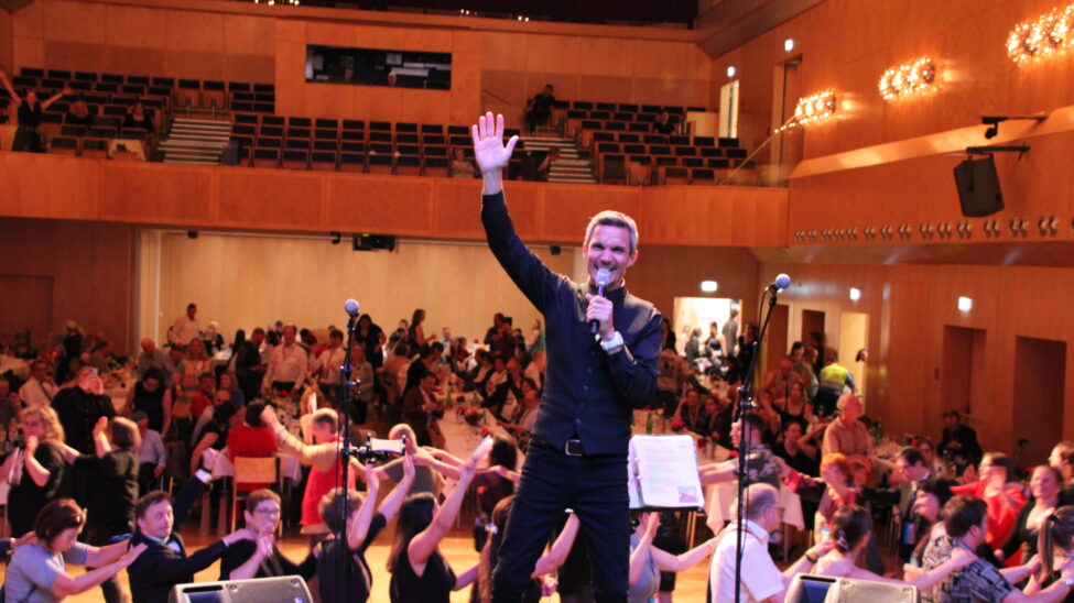 Ein Moderator auf einer Bühne lächelt in die Menge, hebt eine Hand zum Gruß und hält ein Mikrofon. Im Hintergrund sitzen und tanzen viele Gäste in einem festlich beleuchteten Saal, während auf der Bühne Mikrofonständer und Notenständer zu sehen sind.