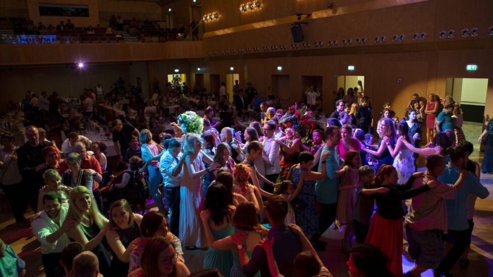 „Große Menschenmenge feiert gemeinsam in einem Saal, viele tanzen in einer langen Polonaise. Die Stimmung ist fröhlich, buntes Licht beleuchtet die Tanzenden, während andere am Rand sitzen oder zuschauen.“
