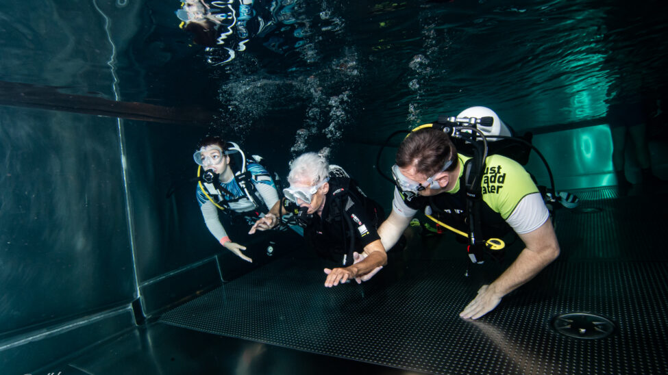 Drei Personen tauchen gemeinsam in einem klaren Becken. In der Mitte bewegt sich eine ältere Person mit Tauchmaske und Atemgerät, links und rechts begleiten sie zwei erfahrene Taucher und geben Unterstützung. Luftblasen steigen nach oben, während alle ruhig am Beckenboden entlanggleiten.