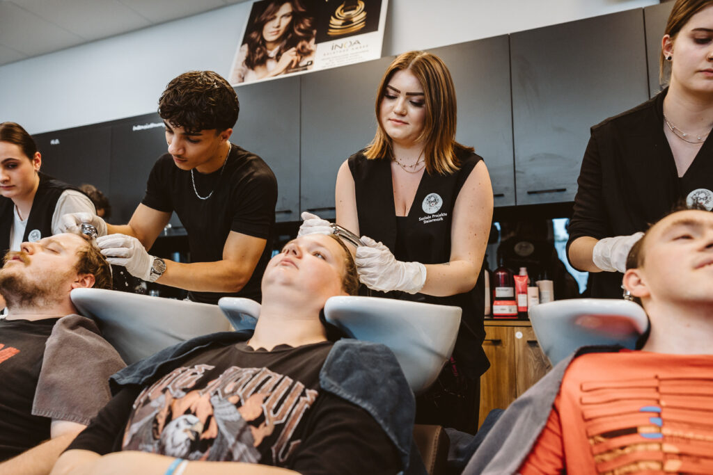 Mehrere Menschen sitzen an Waschbecken in einem Friseursalon, während auszubildende Friseurinnen und Friseure ihnen die Haare waschen. Die Auszubildenden tragen schwarze Arbeitskleidung und Handschuhe, arbeiten konzentriert und wirken aufmerksam. Im Hintergrund stehen weitere Personen und Arbeitsmaterial.