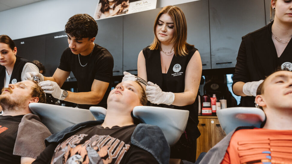 Mehrere Menschen sitzen an Waschbecken in einem Friseursalon, während auszubildende Friseurinnen und Friseure ihnen die Haare waschen. Die Auszubildenden tragen schwarze Arbeitskleidung und Handschuhe, arbeiten konzentriert und wirken aufmerksam. Im Hintergrund stehen weitere Personen und Arbeitsmaterial.