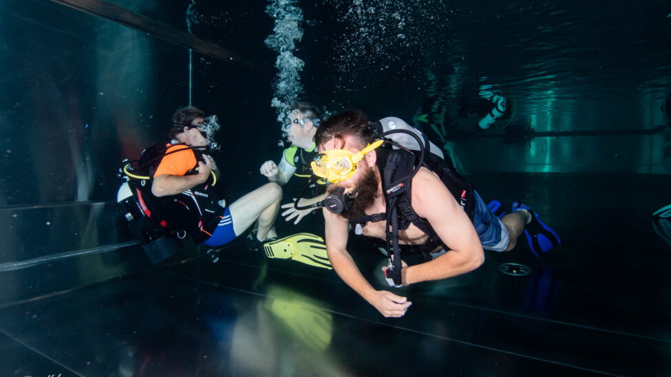 Drei Taucher befinden sich unter Wasser in einem Becken und absolvieren gemeinsam eine Tauchübung. Mit Tauchflaschen, Masken und Flossen bewegen sie sich kontrolliert durch das leicht beleuchtete Wasser, während aus ihren Atemreglern Luftblasen aufsteigen. Die Szene zeigt konzentrierte Zusammenarbeit und sicheres Tauchen in ruhiger Umgebung.