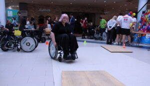 Eine Frau im Rollstuhl fährt konzentriert über einen kleinen Hindernisparcours in einem Einkaufszentrum. Hinter ihr stehen weitere Teilnehmende, einige ebenfalls im Rollstuhl, und verfolgen das Geschehen. Rundherum informieren Stände und Besucherinnen und Besucher über barrierefreie Mobilität.