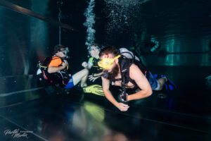 Drei Taucher befinden sich unter Wasser in einem Becken und absolvieren gemeinsam eine Tauchübung. Mit Tauchflaschen, Masken und Flossen bewegen sie sich kontrolliert durch das leicht beleuchtete Wasser, während aus ihren Atemreglern Luftblasen aufsteigen. Die Szene zeigt konzentrierte Zusammenarbeit und sicheres Tauchen in ruhiger Umgebung.