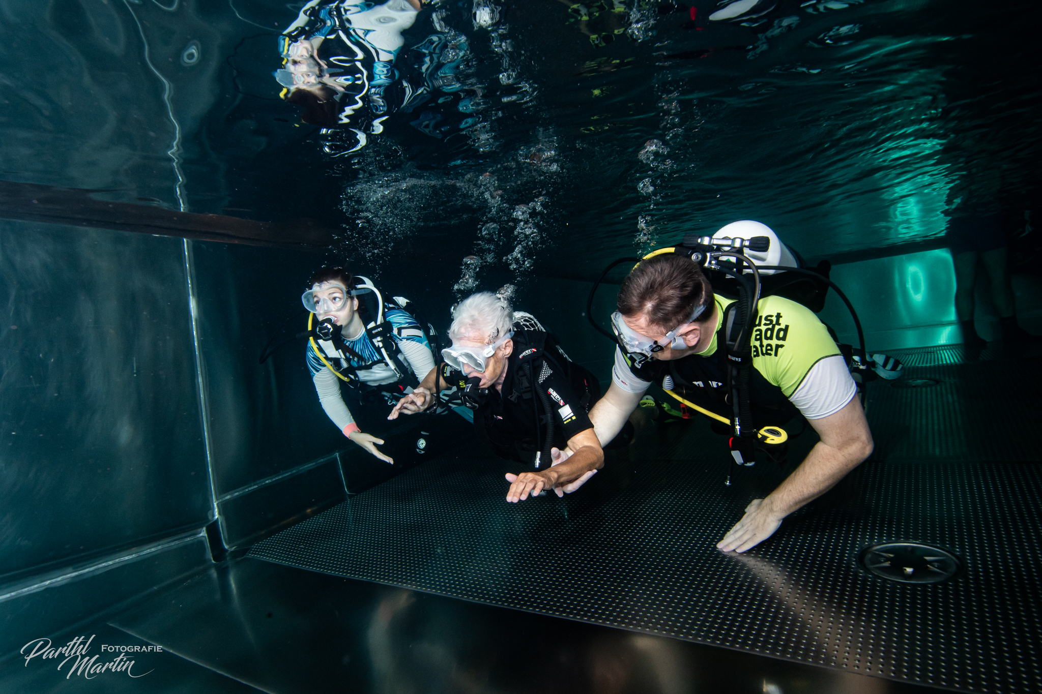 Drei Personen mit Taucherausrüstung befinden sich in einem Unterwasserbecken. In der Mitte wird eine ältere Person von zwei Begleitpersonen links und rechts unterstützt, während alle ruhig über den Beckenboden gleiten. Luftblasen steigen auf, die Szene wirkt wie ein betreutes Tauchtraining in einem Indoor-Tank.